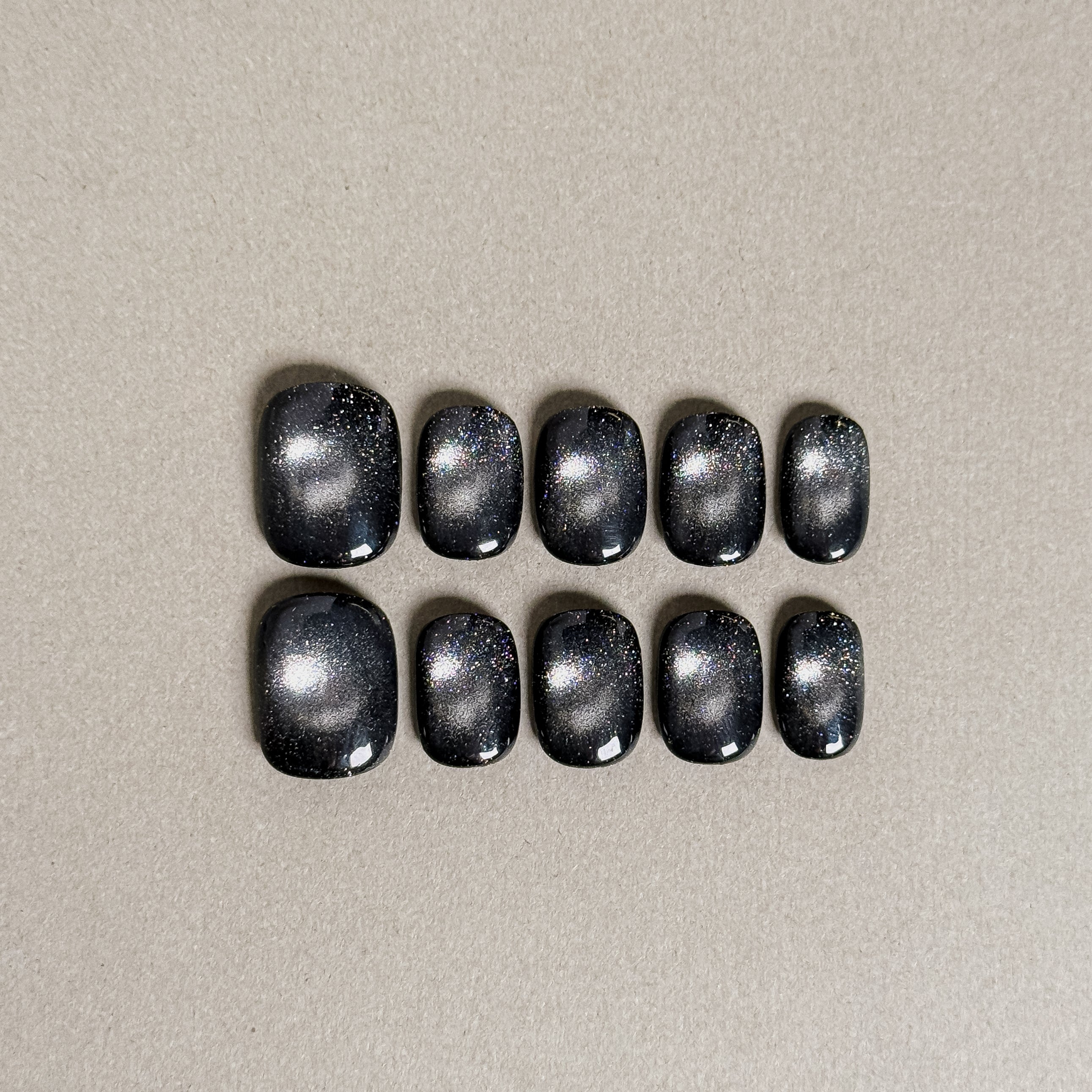 Two rows of MIBLAB's dark gel polish cat eye press-on toenails, showcasing a mesmerizing deep blue-black hue with a shimmering cat eye effect, arranged against a neutral gray background to highlight the unique texture and depth of the polish, with a gradient of sizes from large to small for a perfect fit.