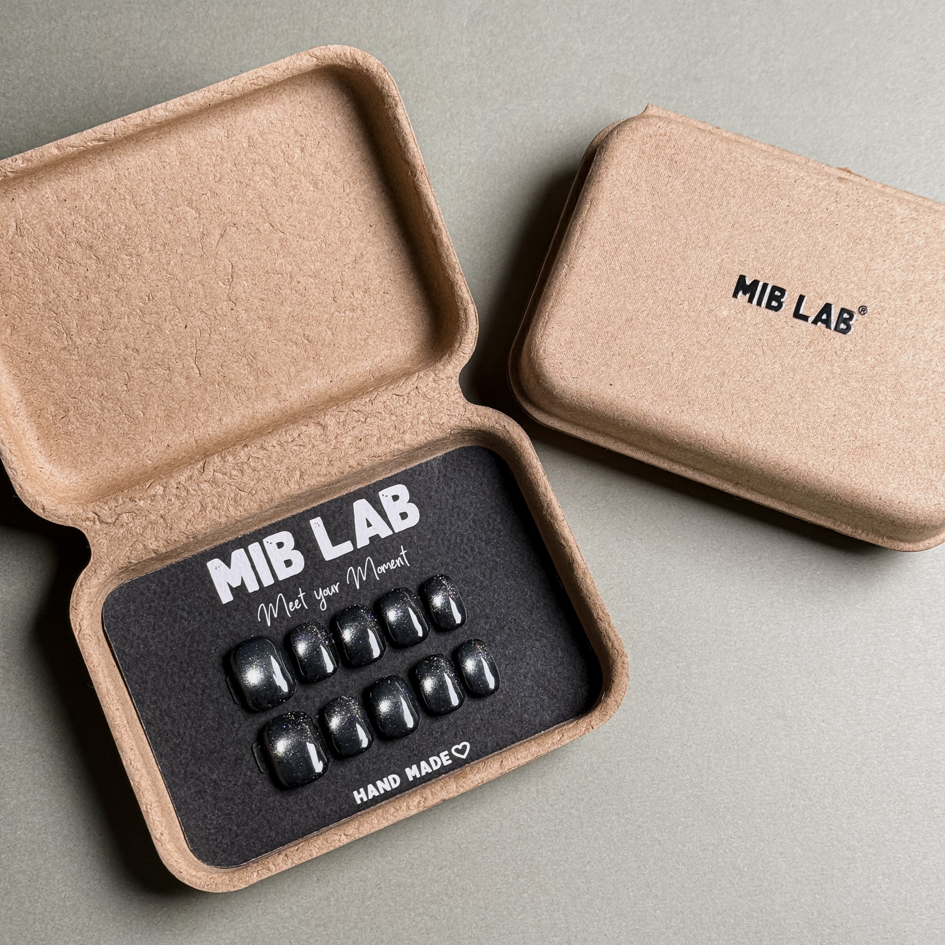 MIBLAB brand press-on toenail set featuring dark gel polish cat eye toenails with a shimmering blue-black cat eye effect, packaged in an eco-friendly recycled paper box with the brand name "MIB LAB" and the slogan "Meet your Moment" displayed, along with the "HAND MADE ♡" inscription, presented against a neutral gray background.