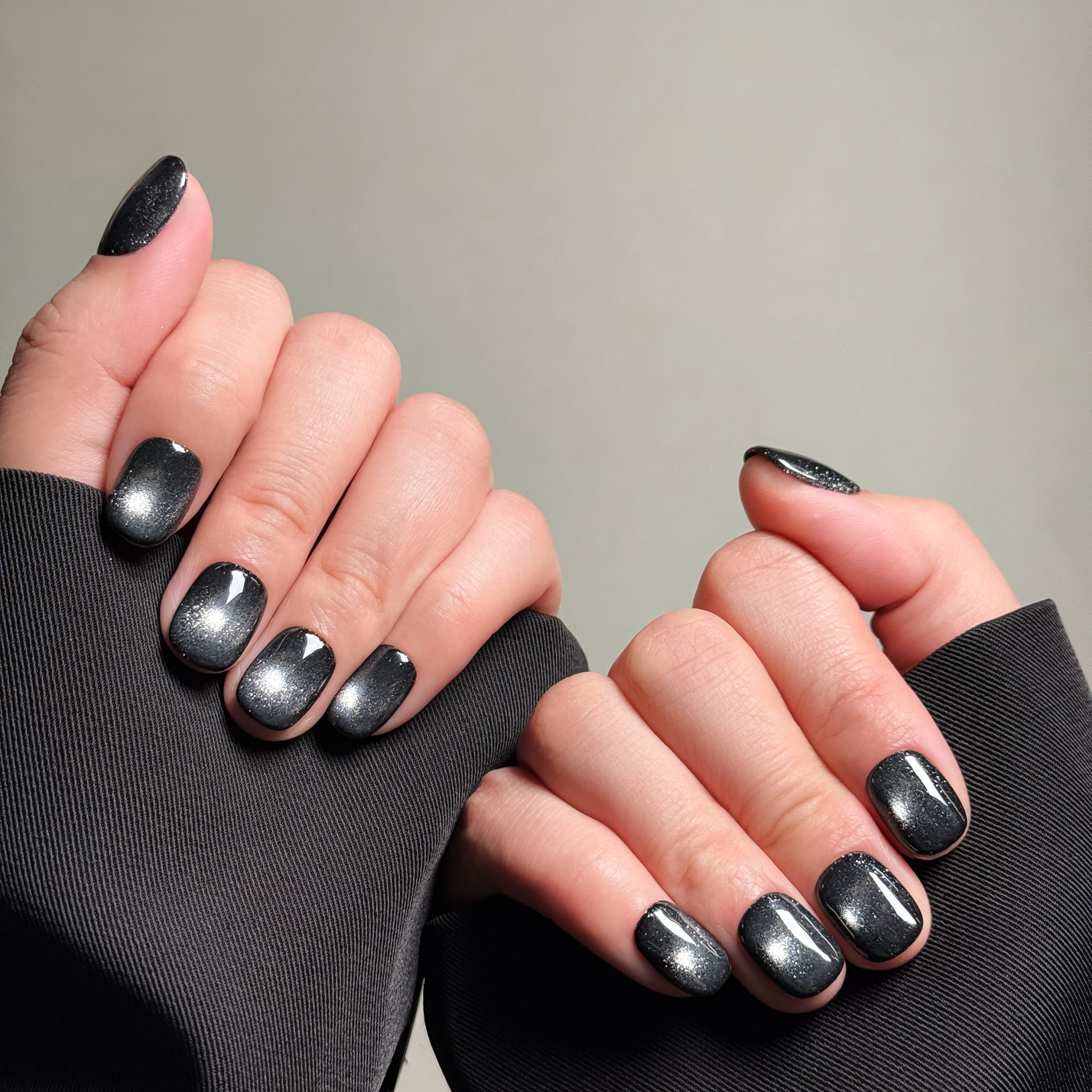 Close-up shot of hands wearing MIBLAB's silver gel polish cat eye press-on nails, showcasing a mesmerizing metallic silver hue with a shimmering cat eye effect, presented against a neutral gray background and a black fabric texture, highlighting the unique texture and depth of the polish on both short and long nail shapes.