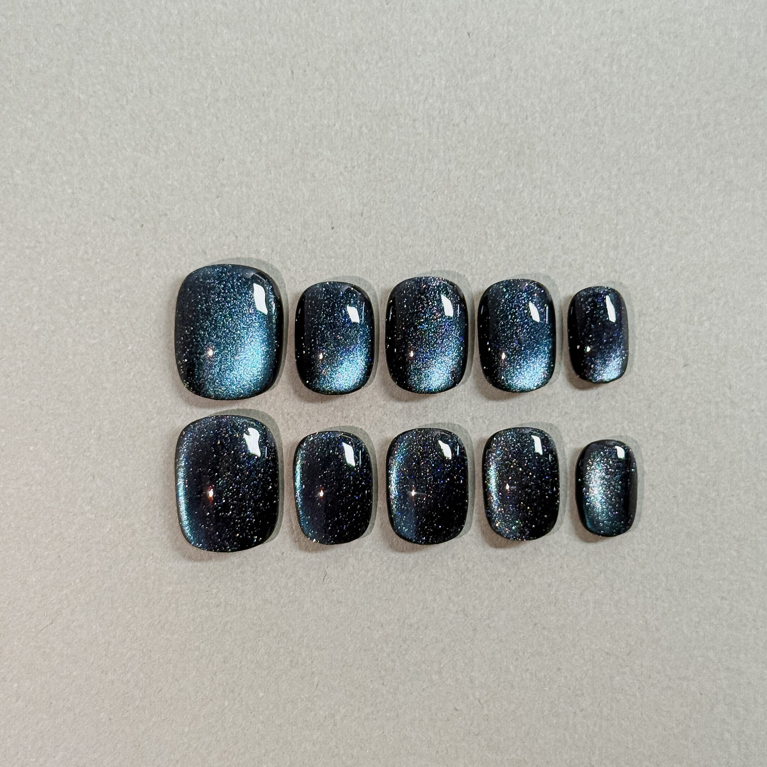 Two rows of MIBLAB's dark gel polish cat eye press-on toenails, showcasing a mesmerizing deep blue-black hue with a shimmering cat eye effect, arranged against a neutral gray background to highlight the unique texture and depth of the polish, with a gradient of sizes from large to small for a perfect fit.
