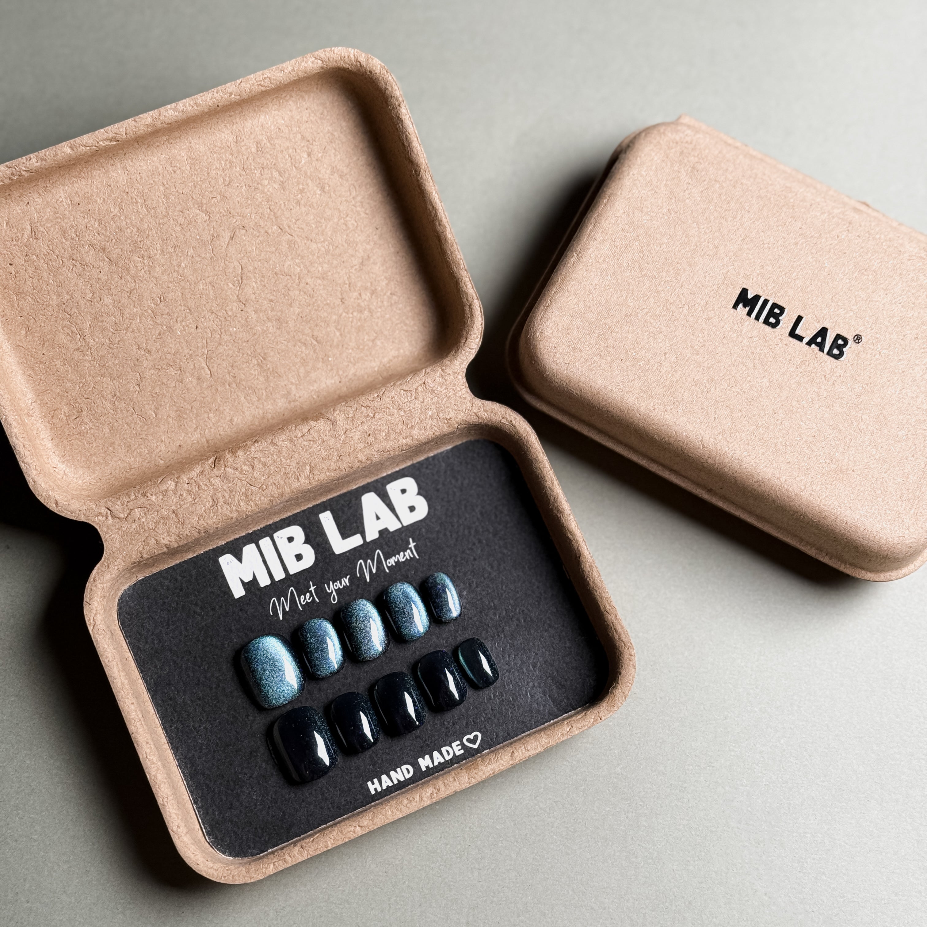 MIBLAB brand press-on toenail set featuring dark gel polish cat eye toenails with a shimmering blue-black cat eye effect, packaged in an eco-friendly recycled paper box with the brand name "MIB LAB" and the slogan "Meet your Moment" displayed, along with the "HAND MADE ♡" inscription, presented against a neutral gray background.