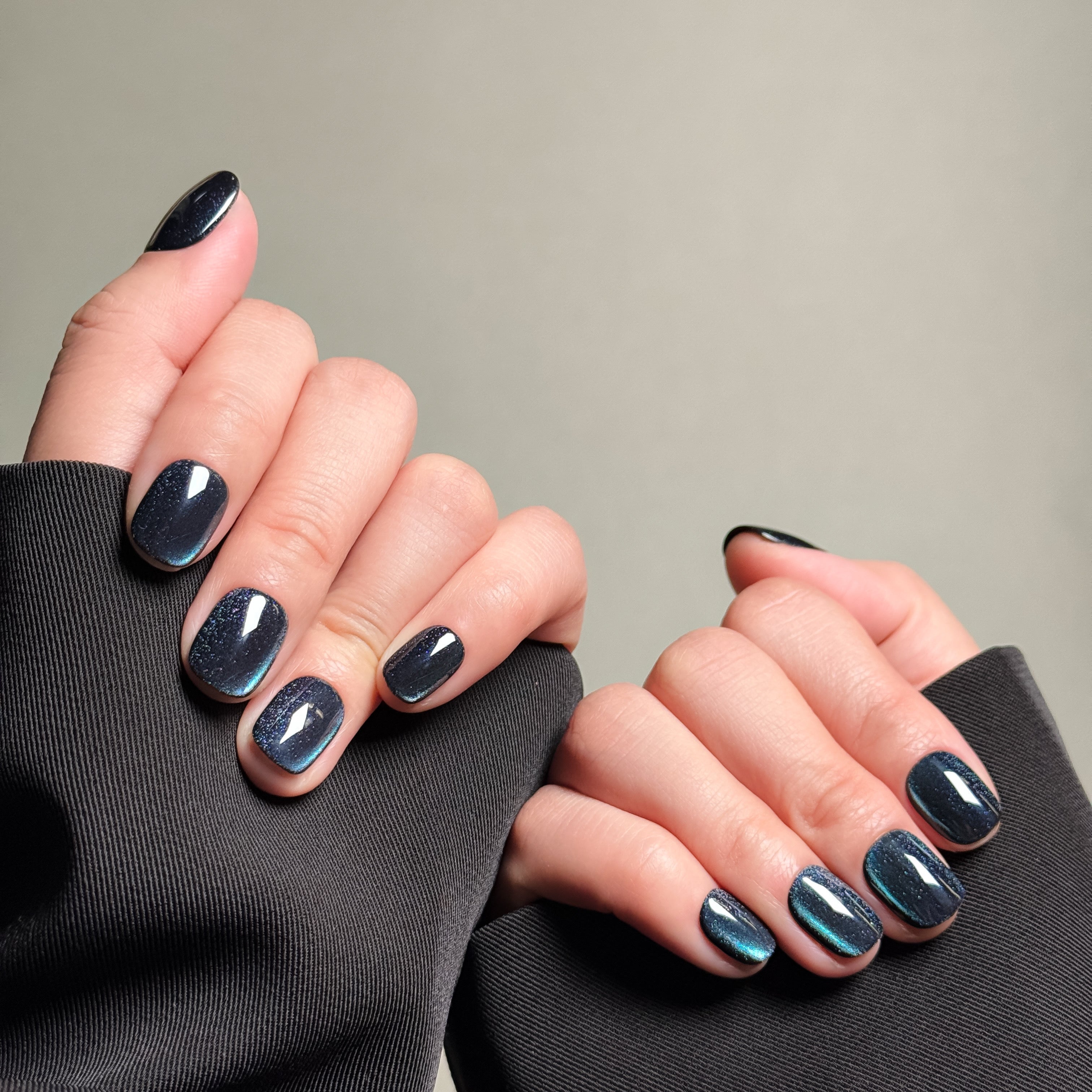 Close-up shot of hands wearing MIBLAB's dark gel polish cat eye press-on nails, showcasing a mesmerizing deep blue-black hue with a shimmering cat eye effect, presented against a neutral gray background and a black fabric texture, highlighting the unique texture and depth of the polish on both short and long nail shapes.