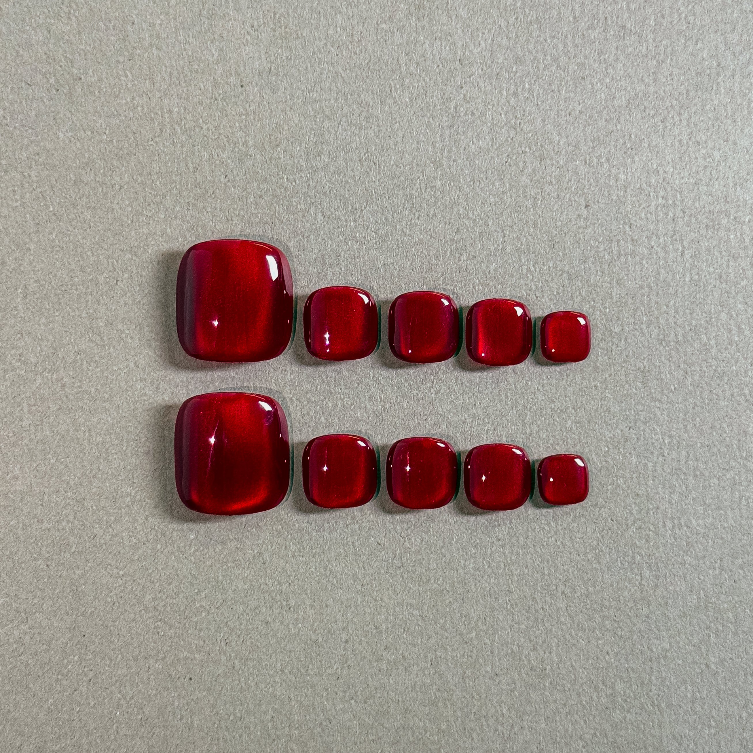 Two rows of MIBLAB's vibrant red press-on toenails, featuring a glossy, gem-like finish reminiscent of rubies, arranged against a neutral gray background, showcasing a gradient of sizes from large to small for a perfect fit, highlighting the luxurious and polished look of the pedicure set.