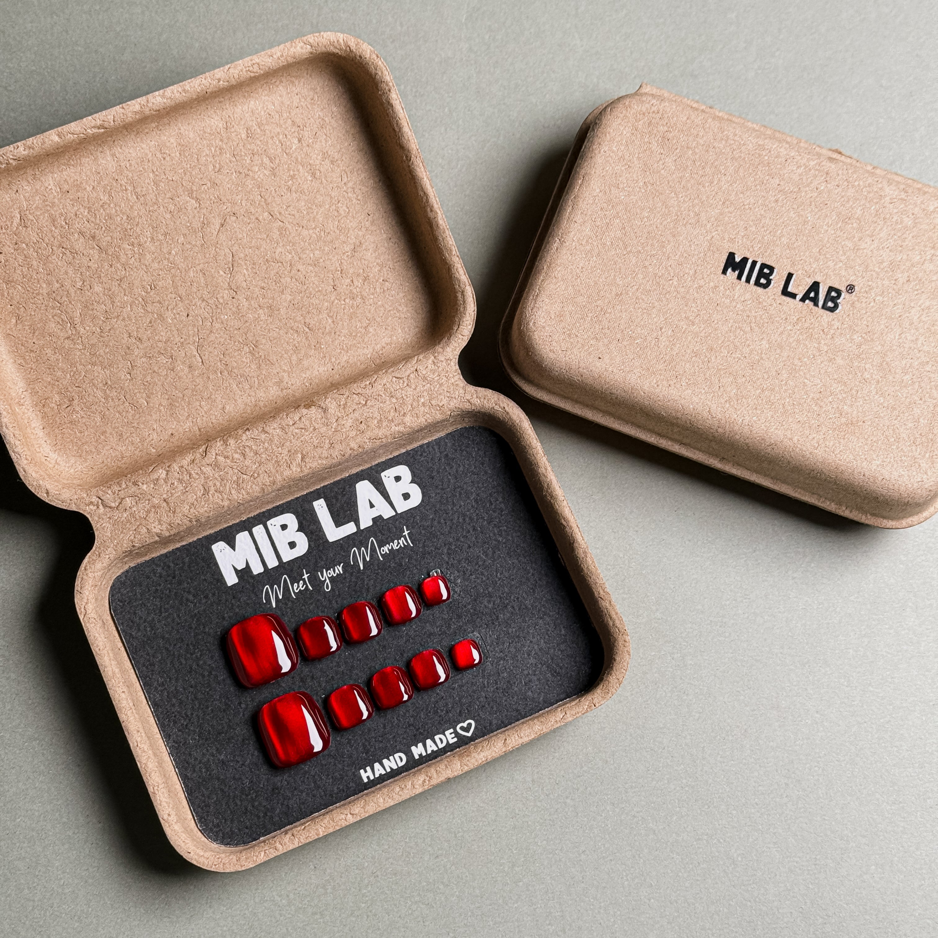 MIBLAB brand press-on toenail set featuring vibrant red, glossy toenails with a gem-like finish, packaged in an eco-friendly recycled paper box with the brand name "MIB LAB" and the slogan "Meet your Moment" displayed, along with the "HAND MADE ♡" inscription, presented against a neutral gray background.