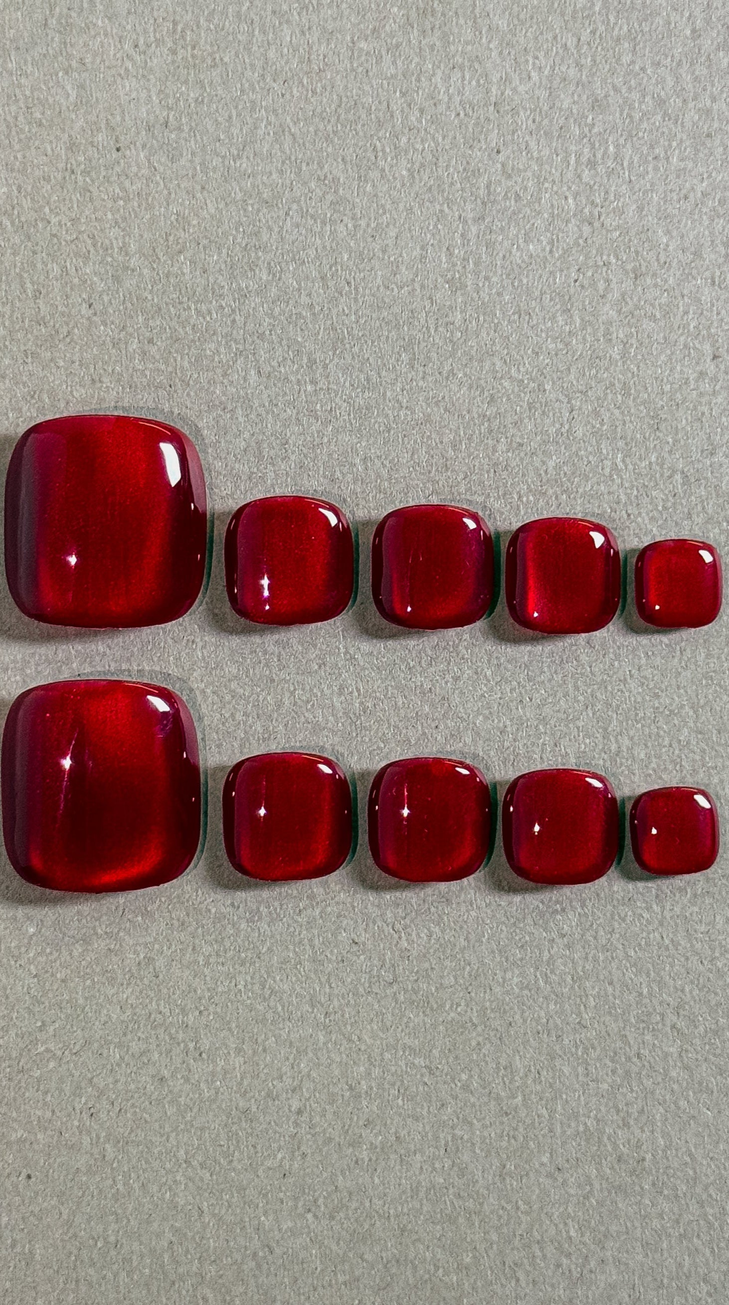 Two rows of MIBLAB's vibrant red press-on toenails, featuring a glossy, gem-like finish reminiscent of rubies, arranged against a neutral gray background, showcasing a gradient of sizes from large to small for a perfect fit, highlighting the luxurious and polished look of the pedicure set.