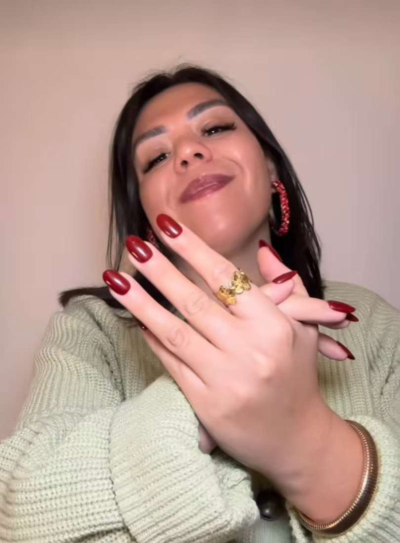 Cliente souriante montre ses press-on nails rouge bordeaux, mains croisées avec bagues, pull beige et boucles d’oreilles assorties.