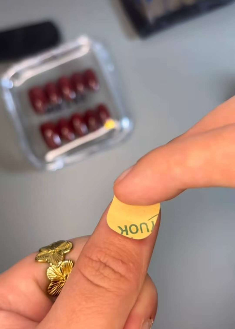 Gros plan : une main avec une bague papillon pose un sticker adhésif sur l’ongle du pouce, avec une boîte de press-on nails rouges floue en arrière-plan.