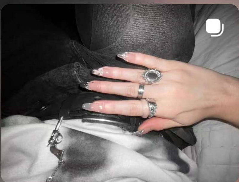 Main posée sur un jean avec plusieurs bagues, montrant des ongles longs nude/transparents avec détails brillants (strass) en bout.