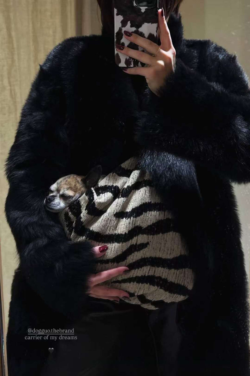 Photo miroir : une personne en manteau en fausse fourrure noire tient un petit chien dans un sac zébré, avec des ongles rouge bordeaux visibles.