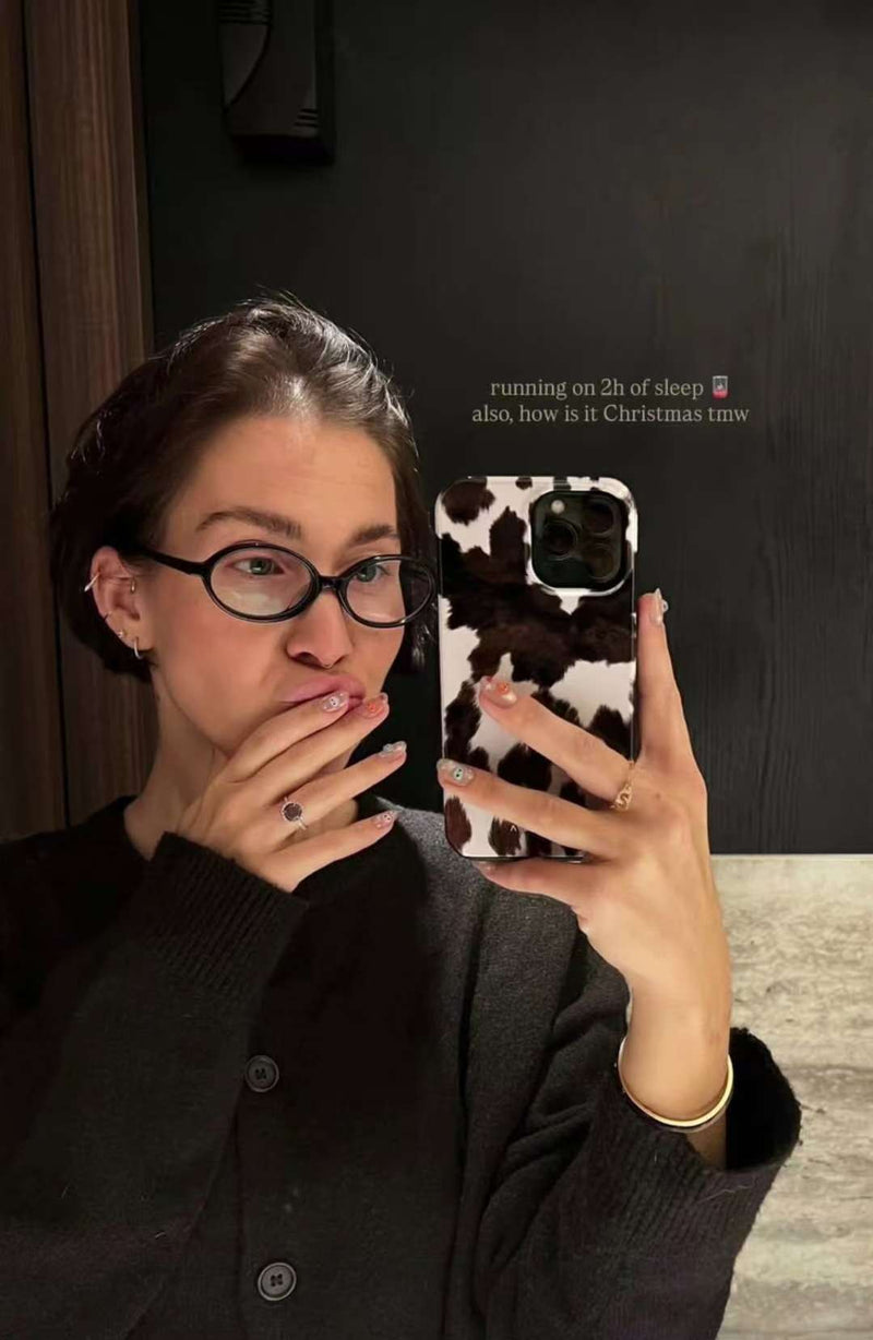 Selfie miroir : personne avec lunettes tient un téléphone à coque “vache” et cache sa bouche avec la main, montrant des press-on nails nude avec petits motifs.
