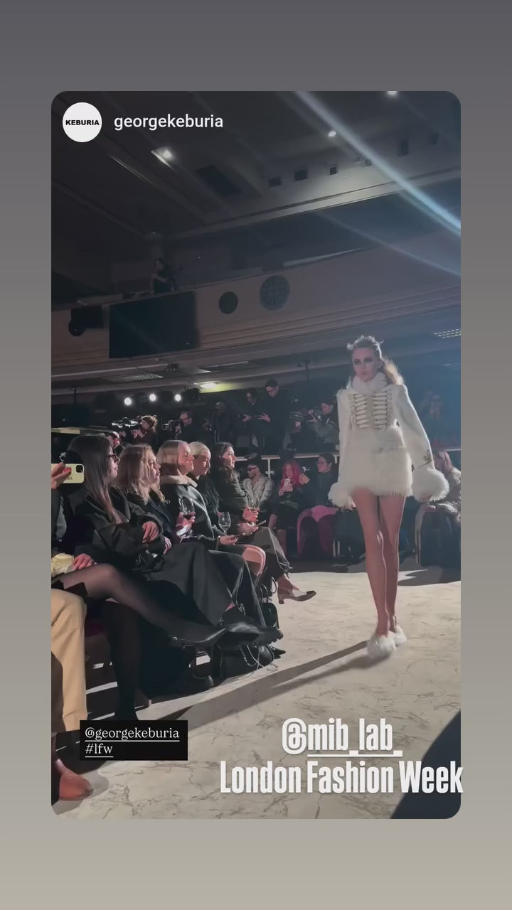 Défilé London Fashion Week : un mannequin en veste blanche effet fourrure défile sur le runway George Keburia, avec le public en arrière-plan.