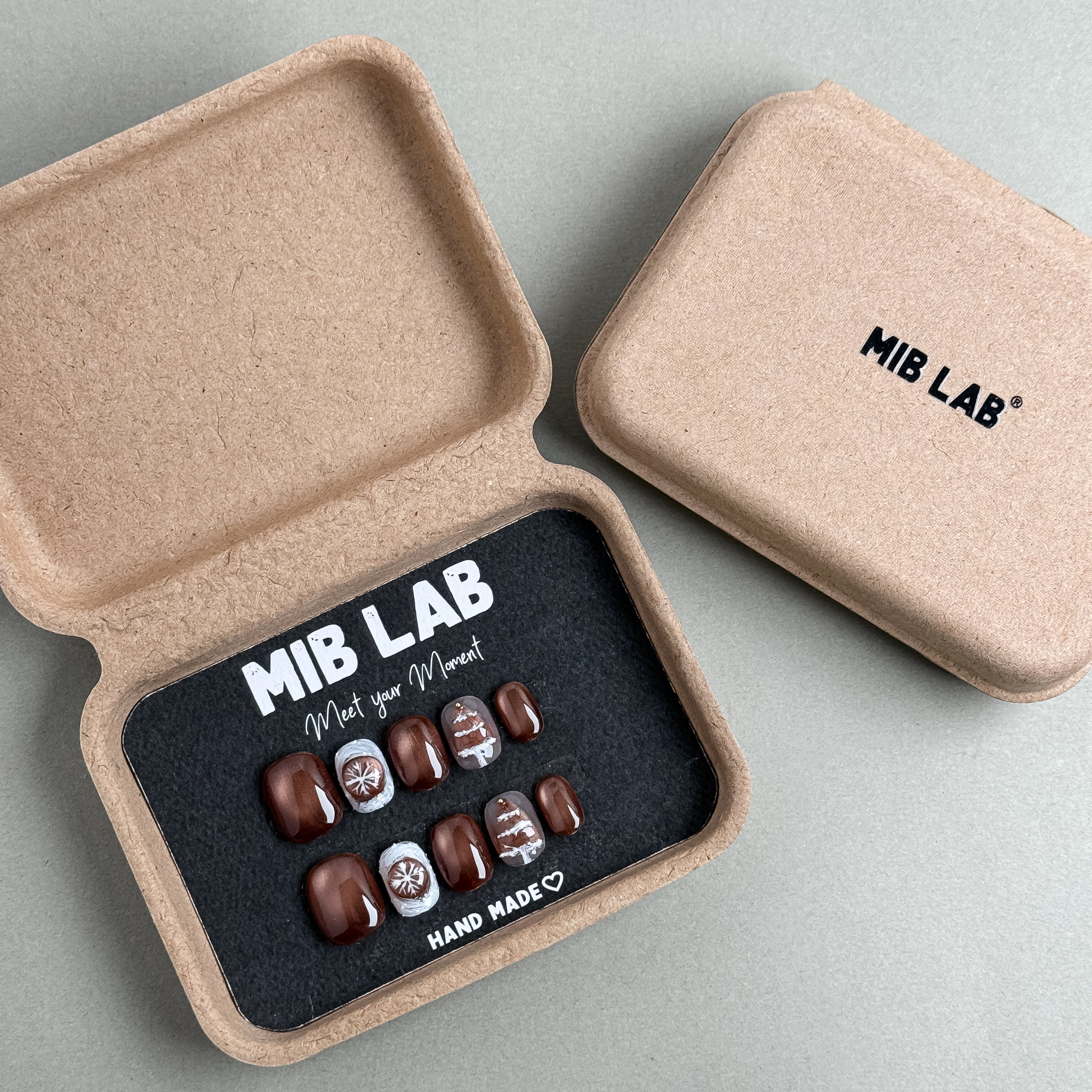 MIBLAB Snow Me press-on nail set in metallic bronze and nude beige with winter snowflake patterns, packaged for boutique display in Geneva.